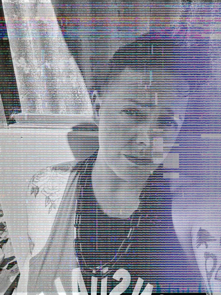 grainy picture of a white person of indeterminate gender with short brown hair wearing a black vest and heavy chain. They are looking at the camera with quizzical eyebrows. They have tattoos of roses on their shoulder and various other objects along their arm.