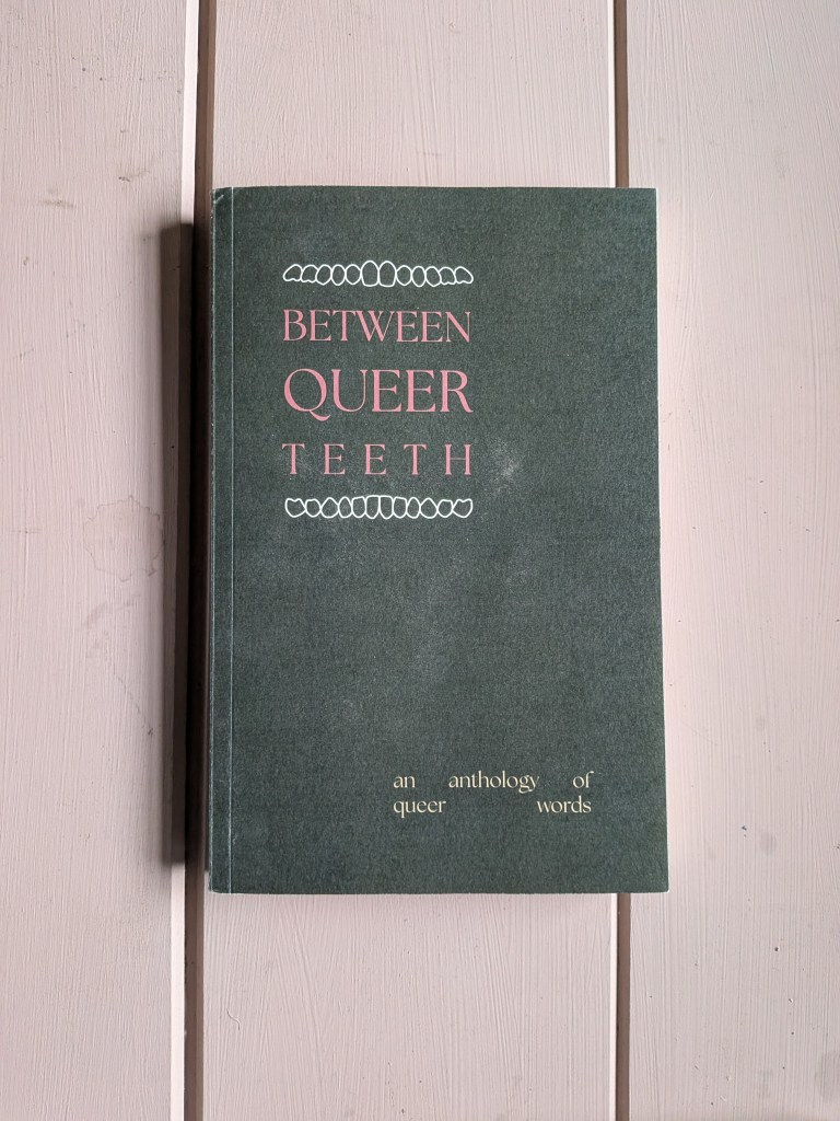 Image of a book against a pink wooden background.
Red text on the olive green cover:
BETWEEN QUEER TEETH
lighter text: an anthology of queer words

Graphics: the title is sandwiched between teeth graphics