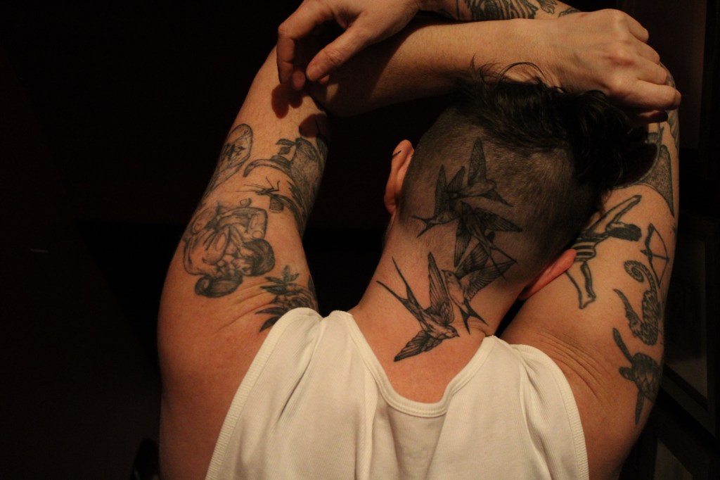 A cryptic headshot of the writer.

Image features the back of a white person's head, their arms raised up, forearms crossed and resting on their head.

They're heavily tattooed, with tattooes of flying swallows on the back of their head, a turtle, seahorse, bull, diver, flowers and queers kissing on the top of their arms, Their hair is shaved into a high fade, there's some short brown hair visible. They have a pierced helix at the top of their left ear. The background is too dark to make any detail out.